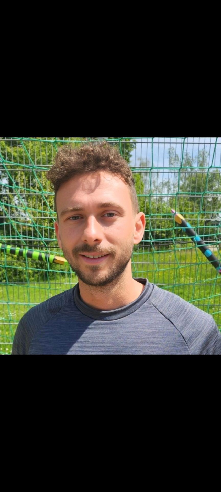 Person standing in front of a fence outdoors on a sunny day.